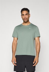 Mees, kes kannab salveerohelist lühikeste varrukatega Nike särk ja musti sportlikke lühikesi pükse, seisab ühetoalist heledat tausta vastas.