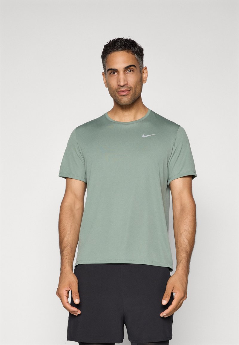 Mees, kes kannab salveerohelist lühikeste varrukatega Nike särk ja musti sportlikke lühikesi pükse, seisab ühetoalist heledat tausta vastas.