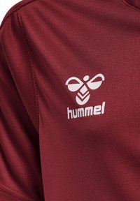 Rød atletisk T-shirt lavet af glat stof med rund halsudskæring. Har et hvidt "hummel" logo og et distinkt bi-emblem på brystet.