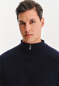 Granatowy sweter na zamek błyskawiczny o ribbowanej teksturze, wysoki kołnierz i mały metalowy zamek. Charakteryzuje się dopasowanym krojem i miękkim wykończeniem.