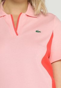 Rosa polo skjorta med detaljer i korall, med en liten grön logotyp och V-halsdesign. Tillverkad av texturerat tyg, kortärmad.
