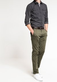 Mörkt mönstrad skjorta med knappar och uppviktärm, i kombination med olivgröna slim-fit byxor och vita sneakers. En brun bälte framhäver outfiten.