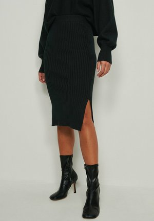 Femme portant une jupe midi en maille côtelée noire avec une fente latérale et des bottines noires à talons sur un fond clair uni.