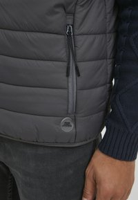 Geacă puffer gri cu cusături orizontale, prevăzută cu un fermoar lateral și un patch cu logo, asortată cu un pulover tricotat texturat de nuanță bleumarin dedesubt.