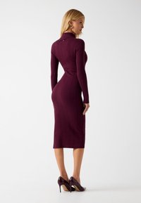 Abito bordeaux in maglia a coste con maniche lunghe e silhouette aderente, caratterizzato da un collo alto e un dettaglio con bottoni sul retro. Indossato con tacchi scuri.