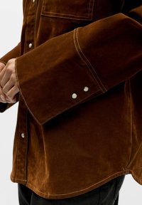 Braunes Baumwollhemd mit einer lockeren Passform, großen Bündchen mit Druckknöpfen, sichtbaren Nähten und einer einzelnen Brusttasche. Glatte Textur.