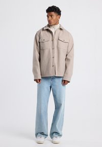 Beige villasekoitus paitajakku, jossa on kaksi läppätaskua, napitus edessä ja rento istuvuus. Yhdistetty vaaleansinsisiin leveälahkeisiin farkkuihin ja valkoisiin lenkkareihin.