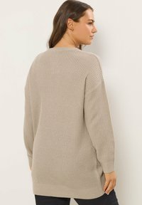 Beżowy, oversize sweter dzianinowy z długimi rękawami, prążkowaną fakturą i luźnym krojem, z bocznymi rozcięciami na końcu dla wygody ruchu.