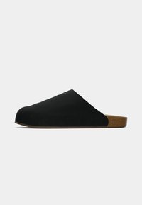 Melni slip-on apavi ar gludu zamšas augšu, noapaļotu purngalu un korķa līdzīgu zoli. Minimālistisks dizains bez redzamām detaļām vai rotājumiem.