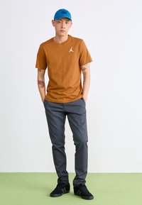 Braun Baumwoll-T-Shirt mit einem kleinen weißen Logo, graue Hose und schwarze Sneakers. Die Kappe ist blau und rundet das legere Outfit ab.