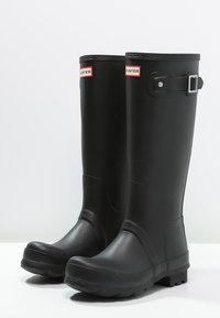 Botas de lluvia de goma negra con acabado brillante, hebilla ajustable y suela texturizada para un mayor agarre. Presenta el logo de Hunter en la parte frontal.