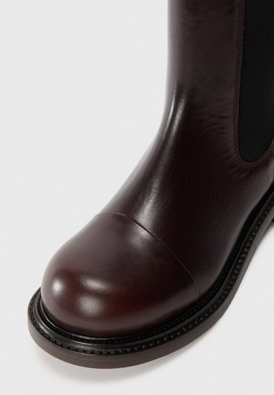 Botină maro din piele, cu vârf rotunjit, panou elastic lateral și talpă texturată. Prezintă o cusătură detaliată de-a lungul marginii tălpii.