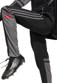 Schwarze und graue Sporthose mit vertikalen grauen Streifen und roten Akzenten. Das Material ist glatt und hat einen taillierten Schnitt, getragen mit schwarzen Fußballschuhen.