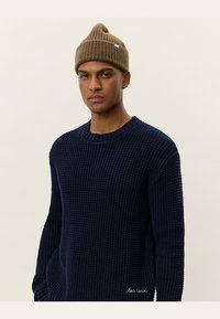 Navy knitted sweater with a textured pattern, round neck, and ribbed cuffs; paired with a brown beanie featuring a peak and logo tag.