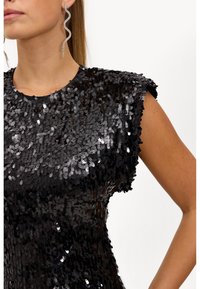 Zwarte top met pailletten, een ronde halslijn en kapmouwtjes, met een gladde textuur en glans van de pailletten; gedragen met spiraalvormige oorbellen.