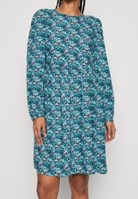 Blommig klänning med långa ärmar, rund halsringning, i blå och gröna nyanser, med en samlad midja och en svängig silhuett.