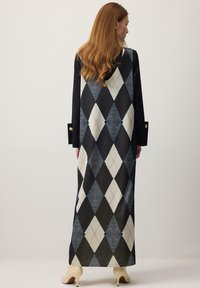 Hosszú vörös hajú nő, hosszú fekete ruhában, amelyen nagy fehér és kék kockás minta látható, fehér falnak háttal áll, bézs magassarkú látható.