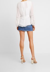 Weiße durchscheinende Bluse mit Spitzenverzierungen, gerafftem Saum und langen Ärmeln, kombiniert mit einem blauen Jeans-Minirock und beigen Sandalen mit klobigem Absatz.