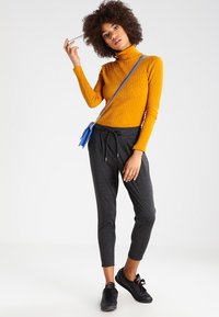 Pletený hořčicový rolák, černé kalhoty se šňůrkou a černé tenisky. Malá modrá crossbody taška, modelka stojí v póze s rukou ve vlasech.