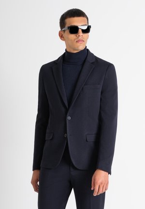Blazer blu navy con due tasche anteriori, rever a forma di V e chiusura con bottone, abbinato a un dolcevita blu navy a costine e occhiali da sole neri.