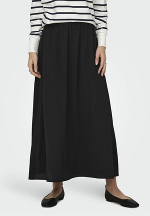 Femme portant une longue jupe noire, des ballerines noires et un haut blanc à manches longues avec des rayures horizontales noires, debout sur un fond uni.