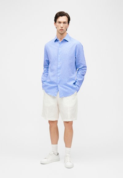 Άνδρας που φοράει ανοιχτό γαλάζιο πουκάμισο με μακριά μανίκια και κουμπιά, λευκά σορτς μέχρι το γόνατο, λευκές κάλτσες και λευκά αθλητικά παπούτσια, στέκεται με τα χέρια στις τσέπες.