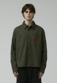 Olive green button-up shirt in a soft fabric, featuring two chest pockets and a red embroidered logo on the left side.
