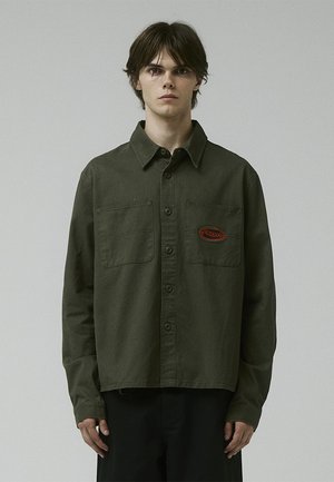 Olive green button-up shirt in a soft fabric, featuring two chest pockets and a red embroidered logo on the left side.