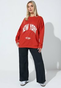 Felpa rossa con la scritta "NEW YORK" stampata in bianco, caratterizzata da una vestibilità comoda e scollo rotondo. Abbinata a pantaloni neri a gamba larga e sneakers.