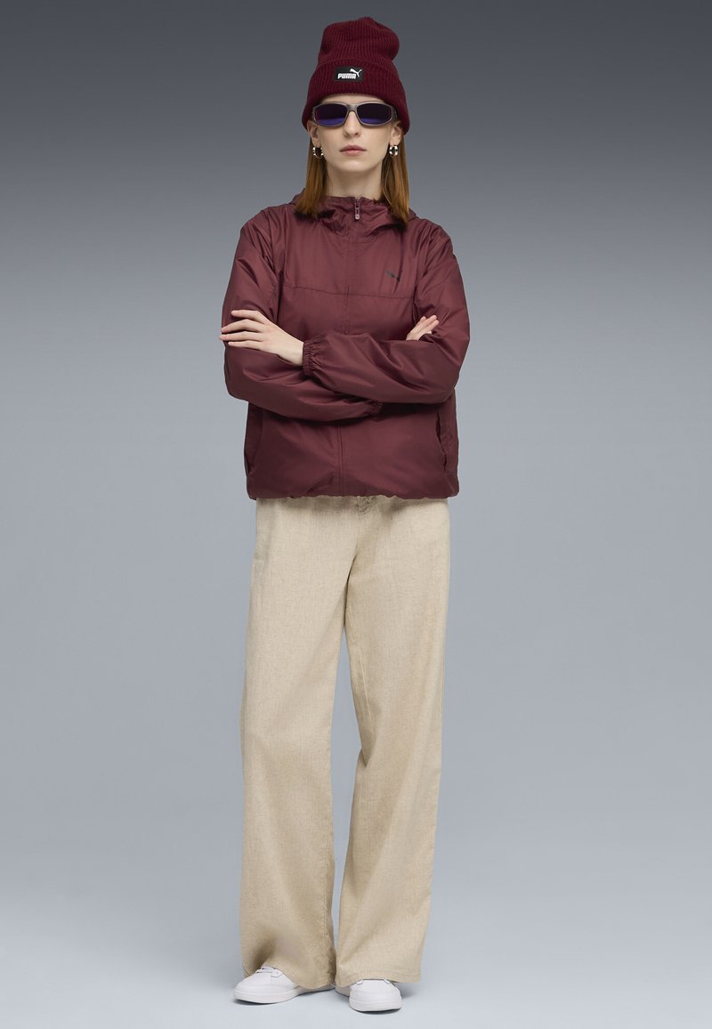Bordeauxfarbene leichte Jacke mit Kapuze und Kordelzug, kombiniert mit cremefarbenen weiten Hosen und weißen Schuhen. Dazu gehören ein bordeauxfarbener Beanie und Sonnenbrille.