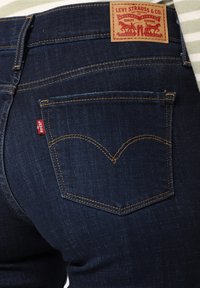 Jean en denim bleu foncé avec une texture subtile, doté de poches arrière classiques et d'une étiquette logo rouge. Comprend une pièce en cuir fauve sur la ceinture.
