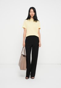 Geel katoenen T-shirt met wit logo, zwarte wijde broek en een beige tote bag. Model draagt zwarte sandalen met open tenen.