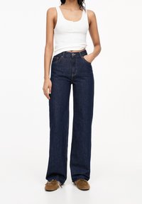 Dark blue jeans with a high waist, wide leg design, and frayed hems. Paired with a white sleeveless top and brown shoes.
