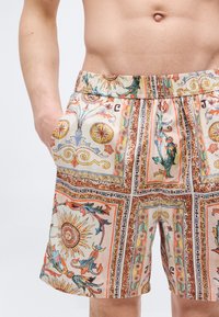 Άνδρας που φοράει πολύχρωμα σορτς με σχέδια από ήλιο, λουλούδια και διακοσμητικά μοτίβα, στέκεται με το ένα χέρι στην τσέπη.