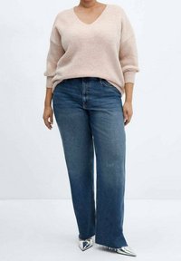 Lichtroze, gebreide trui met een V-hals en een oversized pasvorm, gecombineerd met hoog-waist, wijde blauwe jeans. Zilveren puntige schoenen.