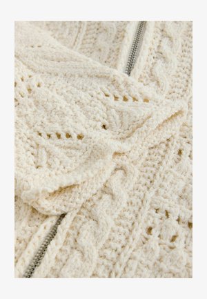 Tekstūruota kreminės spalvos megztinė medžiaga su pintų raštų, su dalinai atidaryta metaline užtrauktuku. Minkštas ir šiltas išvaizda.