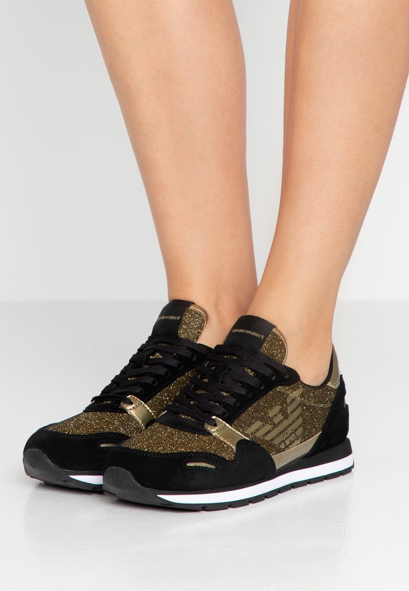 Jambes portant des baskets scintillantes noires et dorées avec des semelles blanches, des lacets noirs et un logo ailé sur le côté.
