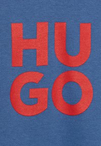 La tela de algodón azul presenta letras en bloque rojas y en negrita que dicen "HUGO". La textura es acanalada, lo que enfatiza la impresión.