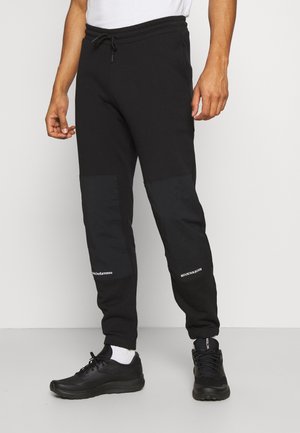Homme portant un pantalon de jogging noir avec des renforts aux genoux et des baskets noires, debout devant un fond clair uni.