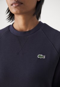 Marineblå hettegenser med teksturert stoff, rund hals og en liten grønn og hvit brodert krokodillelogo på brystet.