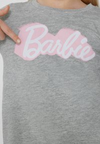 Šedé bavlněné tričko s výrazným růžovým a bílým logem "Barbie" umístěným na hrudi. Materiál vypadá měkce a hladce.