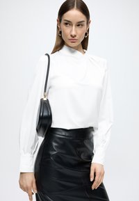 Bluza albă cu mâneci lungi, guler înalt și detalii fronsate, asortată cu o fustă conică din piele neagră, cu talie înaltă, și o geantă mică neagră.