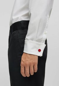 Weißes Langarmhemd mit Knopfmanschetten und einem roten runden Akzent, kombiniert mit schwarzen Hosen. Glatter Stoff, schlanker Schnitt.