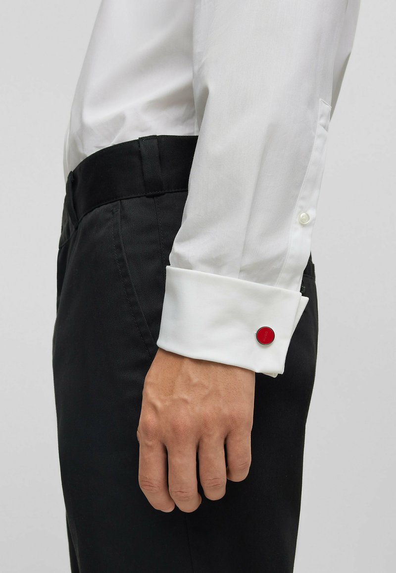 Weißes Langarmhemd mit Knopfmanschetten und einem roten runden Akzent, kombiniert mit schwarzen Hosen. Glatter Stoff, schlanker Schnitt.