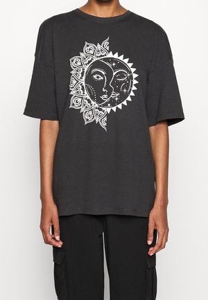 Person wearing a black oversized T-shirt with a white sun and moon face graphic on the chest, paired with black cargo pants.