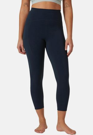Frau in hoch taillierten, dunkelblauen kurzen Leggings und einem hellgrauen Sport-BH, steht barfuß mit entspannten Armen seitlich am Körper.