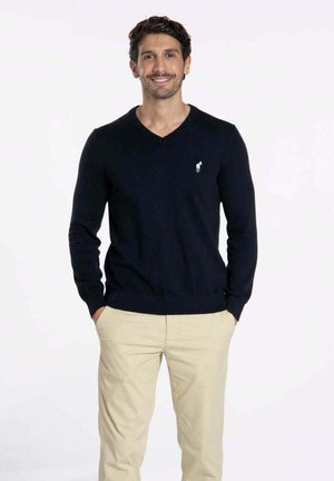 Pull en maille bleu marine avec un col en V, des manches ajustées et un ourlet côtelé, comportant un petit logo blanc sur la poitrine gauche. Associé à un pantalon beige.