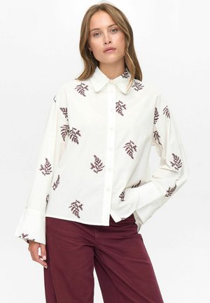 Woman wearing a white button-up blouse with brown leaf patterns and burgundy pants, standing with one hand in pocket against white background.