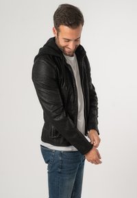 Giacca in pelle nera con cappuccio, dotata di zip frontale, due tasche laterali e finitura texturizzata, indossata sopra una maglietta bianca e jeans.