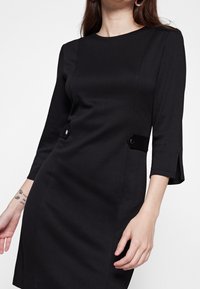 Musta mekko, jossa on kolme neljänneksen hihaa, sivupainikkeet, tyylikäs leikkaus ja hieman sivuhalkioita hihojen helmassa. Sileä kangaspinta.